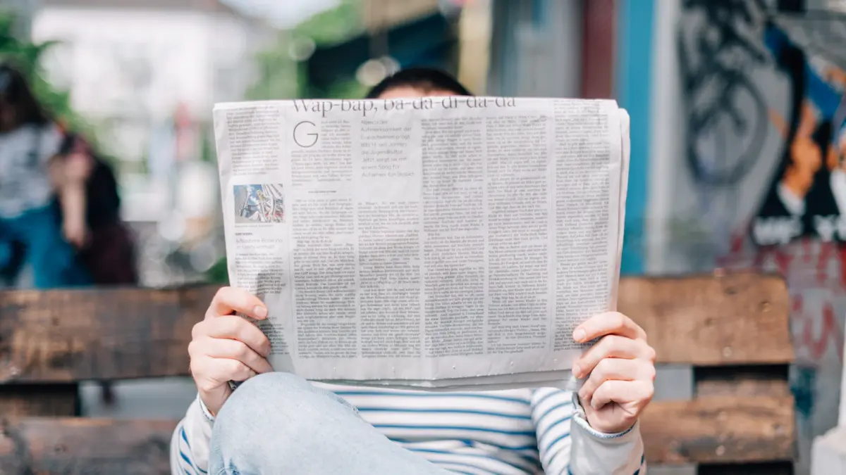 Person reading a newspaper on a wooden bench